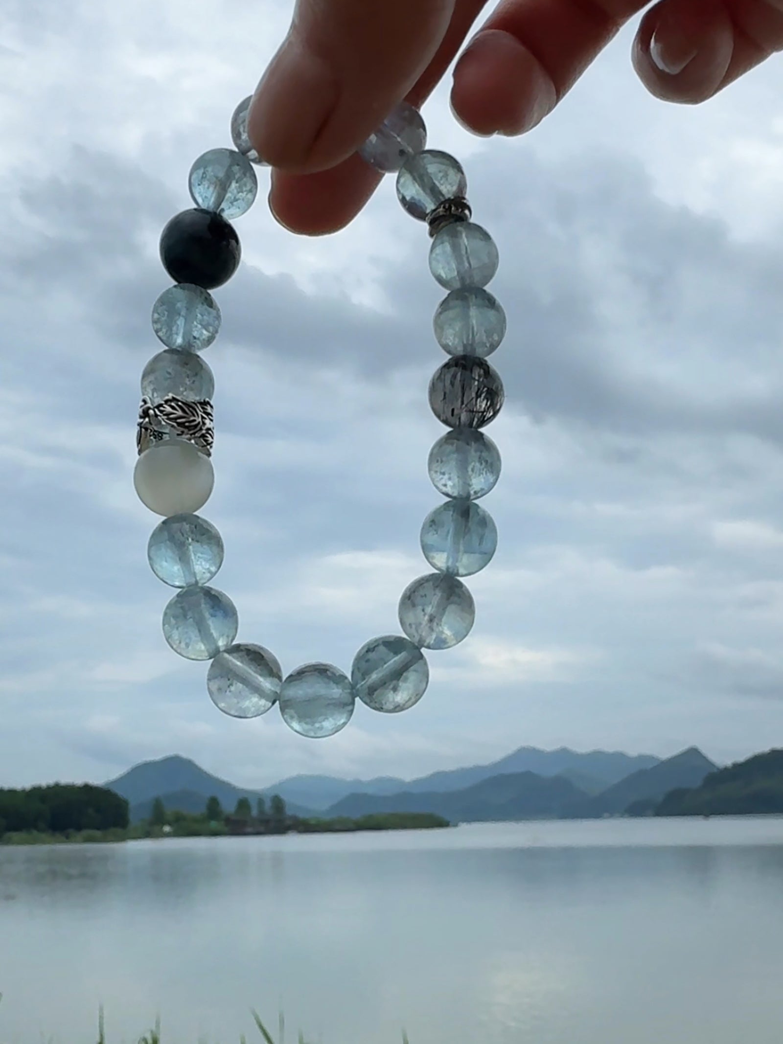 Aquamarine crystal bracelet held beside a quiet mountain lake under cloudy skies, inspired by stillness, calm presence, and Eastern mountain-water aesthetics.”