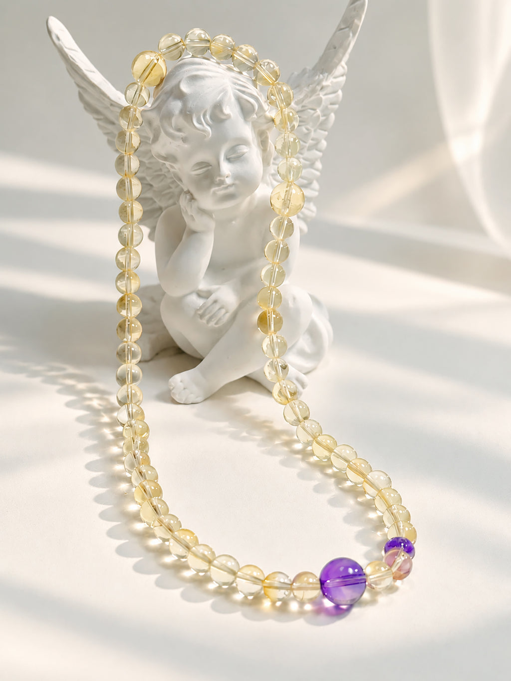Translucent citrine and purple amethyst bracelet elegantly draped over a white angel sculpture in soft natural sunlight.