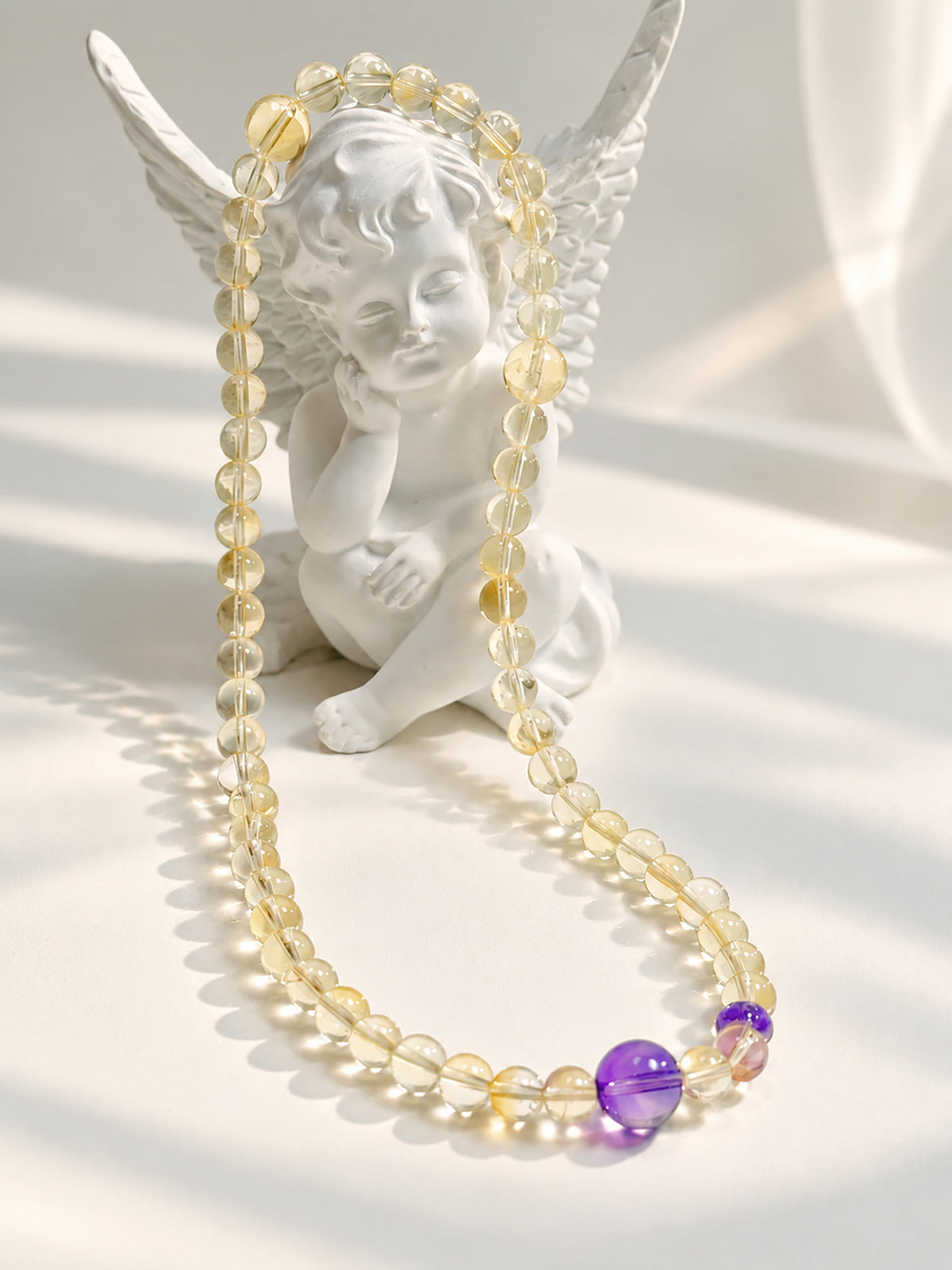Translucent citrine and purple amethyst bracelet elegantly draped over a white angel sculpture in soft natural sunlight.
