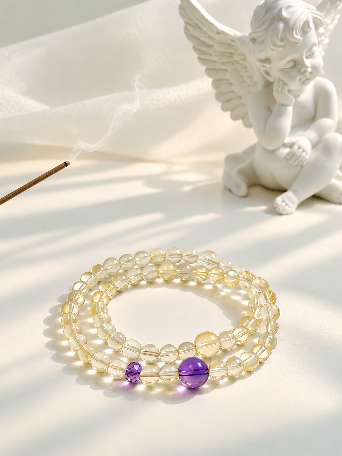Citrine and amethyst double-wrap bracelet arranged beside incense and a white angel sculpture in a softly sunlit setting.